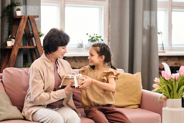 Szczęśliwa dziewczynka w eleganckim stroju casualowym patrząca na swoją uśmiechniętą babcię siedzącą na kanapie i przekazującą jej pudełko z prezentem