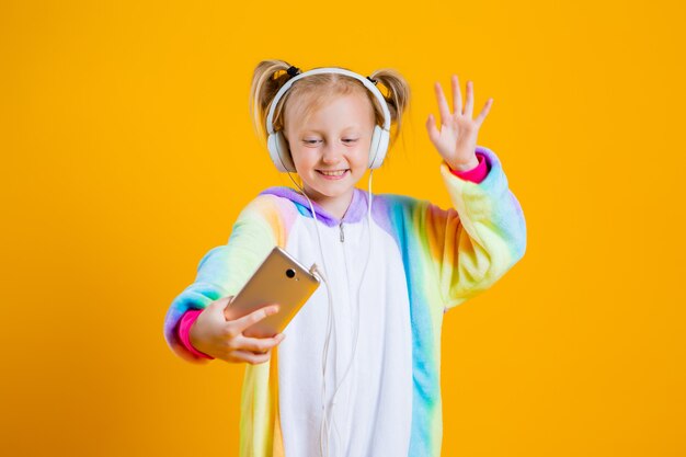 Szczęśliwa dziewczynka w jednorożcu kigurumi słucha muzyki w słuchawkach, trzymając smartfon