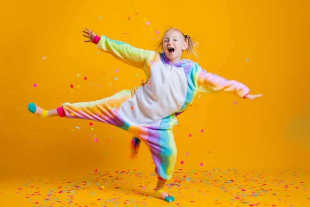 Szczęśliwa dziewczynka w kigurumi jednorożca, taniec na żółtej ścianie wśród wielokolorowych konfetti