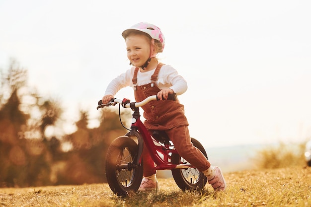 Szczęśliwa dziewczynka w ochronnym kapeluszu jeżdżąca na rowerze na zewnątrz w słoneczny dzień w pobliżu lasu