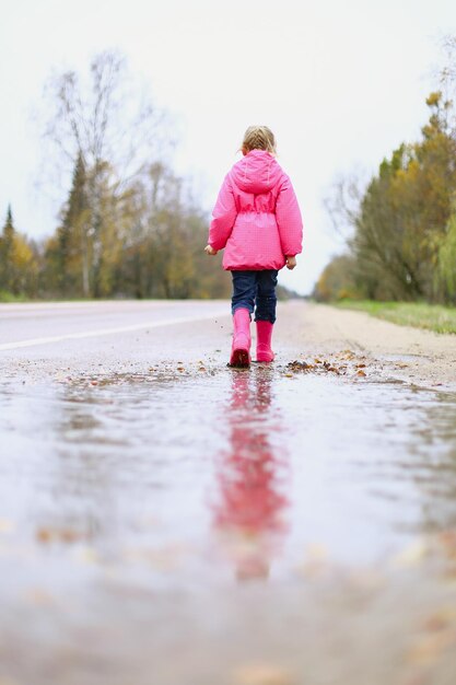 Szczęśliwa dziewczynka w różowej wodoodpornej kurtce kalosze radośnie skacze przez kałuże na ulicy w deszczową pogodę Wiosna jesień Dziecięca zabawa na świeżym powietrzu po deszczu Rekreacja na świeżym powietrzu