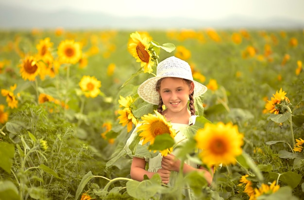 szczęśliwa dziewczynka w zielonej sukience i białym kapeluszu stoi w warkoczu z uśmiechniętymi żółtymi słonecznikami