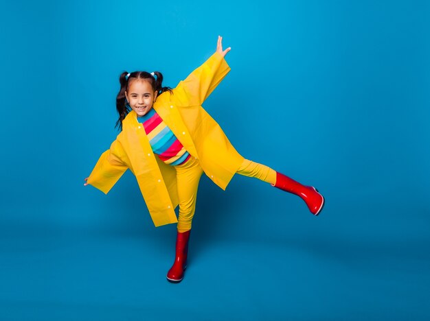 Zdjęcie szczęśliwa dziewczynka w żółtym płaszczu przeciwdeszczowym i czerwonych butach na niebieskim tle.