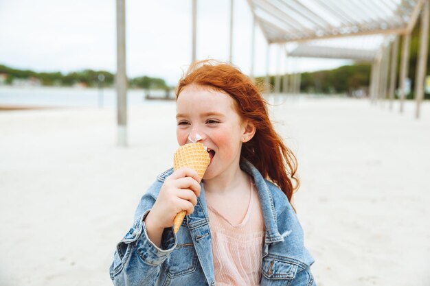 Szczęśliwa dziewczynka z rudymi włosami je lody na plaży w letnie wakacje