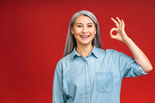 Szczęśliwa elegancka dojrzała starsza kobieta w wieku azjatyckim uśmiechnięta stojąca na białym tle na czerwonym tle Uśmiechnięta pewna siebie wesoła dama w średnim wieku kopia przestrzeń Ok znak