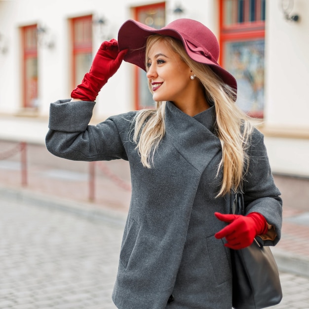 Szczęśliwa elegancka piękna kobieta w kapeluszu z rękawiczkami w szarym płaszczu z czarną torebką na ulicy