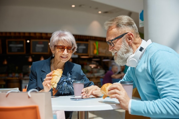Szczęśliwa emerytowana para jedząca croissanty w kawiarni w centrum handlowym