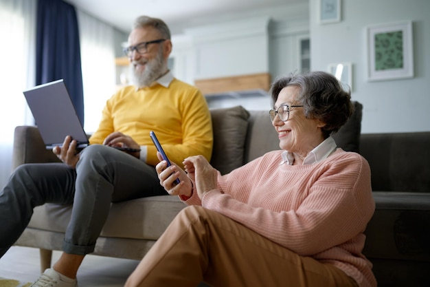 Szczęśliwa emerytowana para korzystająca z cyfrowego gadżetu podczas odpoczynku w salonie w domu. Starsza żona rozmawiająca z przyjaciółmi na smartfonie i starszy mąż przesyłający wiadomości do mediów społecznościowych na laptopie