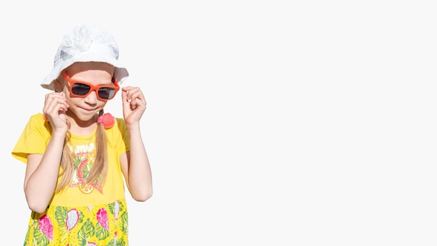 Szczęśliwa emocjonalna dziewczynka w kapeluszowych okularach przeciwsłonecznych i żółtej sukience śmieje się i zakłada okulary przeciwsłoneczne
