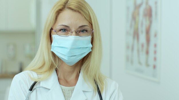 Szczęśliwa europejska kobieta lekarz z okularami w masce medycznej na sobie biały fartuch lekarski i stetoskop, patrząc na kamery. Uśmiechnięta kobieta lekarz pozowanie w biurze szpitala.