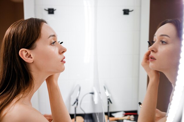 Szczęśliwa glamour dziewczyna patrząc w lustro, stosując tusz do rzęs na rzęsy w łazience Młoda kobieta zakładanie produktu kosmetycznego do makijażu po porannym prysznicu