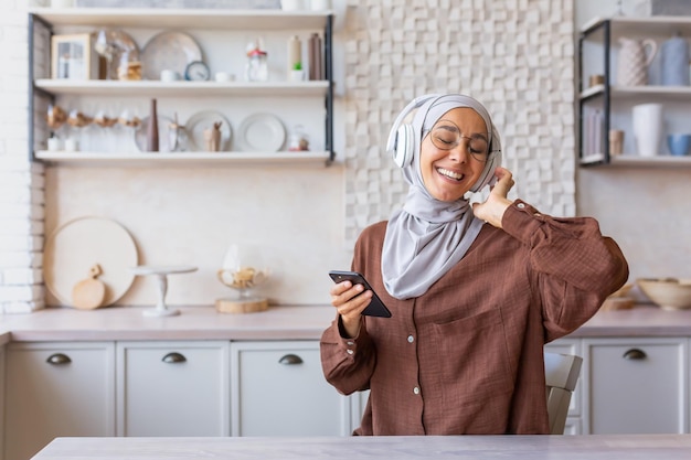 Szczęśliwa i uśmiechnięta kobieta tańczy i śpiewa w domu w kuchni muzułmanki w hidżabie i słuchawkach