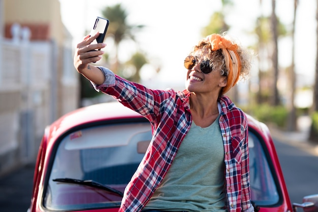 Szczęśliwa i wolna dorosła ładna kobieta uśmiecha się przed swoim samochodem i robi zdjęcie selfie ze smartfonem, ludzie cieszą się podróżami życia i letnimi wakacjami dla jednej pięknej kobiety