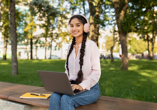 Szczęśliwa indyjska dama relaksuje się z laptopem i słuchawkami w parku, siedząc na ławce i uśmiechając się do kamery