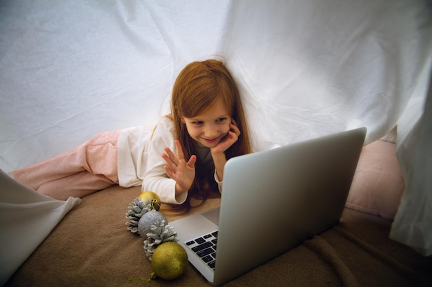 Szczęśliwa kaukaska dziewczynka podczas rozmowy wideo lub wiadomości ze Świętym Mikołajem za pomocą laptopa i urządzeń domowych