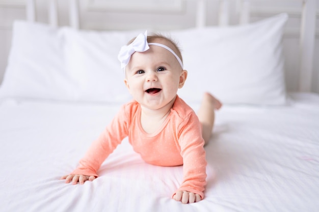 Szczęśliwa kaukaska dziewczynka w różowym body leży na łóżku w domu w sypialni na białej pościeli patrzy w kamerę śmieje się zdrowe dziecko
