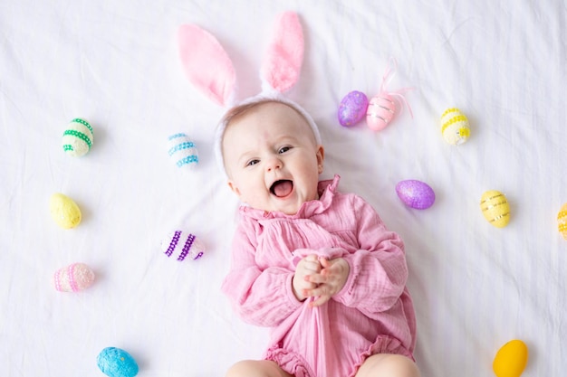 Szczęśliwa kaukaska dziewczynka z uszami królika na głowie leży na plecach na łóżku w domu w sypialni z kolorowymi jajkami wielkanocnymi patrzy w kamerę śmiejąc się Wielkanocne dziecko