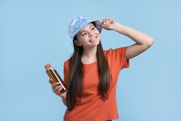 Szczęśliwa kaukaska nastolatka w pomarańczowej koszulce i stylowym niebieskim kapeluszu panama uśmiecha się uroczo, odwraca wzrok, trzymając butelkę z orzeźwiającą lemoniadą odizolowaną na niebieskim tle Koncepcja lata