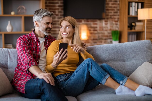 Szczęśliwa kaukaska żona w średnim wieku pokazuje smartfona mężowi, oglądając wideo siedząc na kanapie