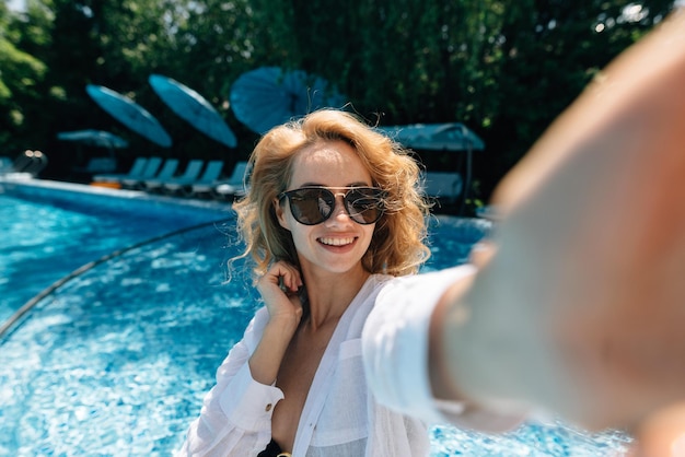 Szczęśliwa kobieta blogerka nagrywa transmisję na żywo w pobliżu basenu w hotelu Dziewczyna w okularach przeciwsłonecznych na wakacjach robi selfie na zbliżenie telefonu komórkowego Wakacje i blogowanie