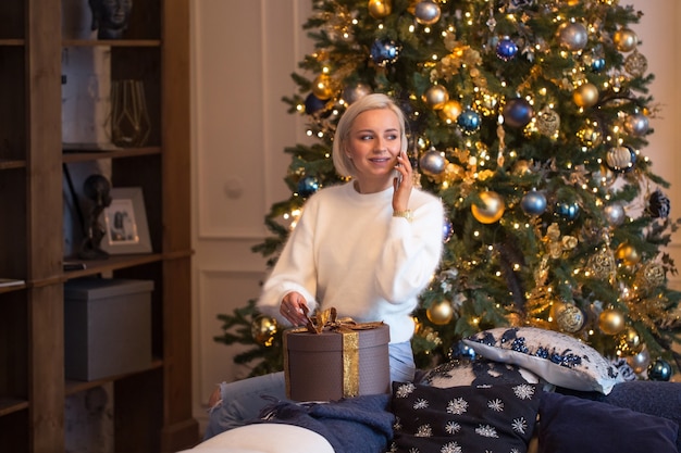 Szczęśliwa kobieta dzwoni do rodziny lub męża wdzięczna za prezent na Boże Narodzenie lub nowy rok siedząc z prezentem w pobliżu choinki w świąteczny poranek. Młoda dziewczyna mówi dziękuję za niespodziankę na ferie zimowe