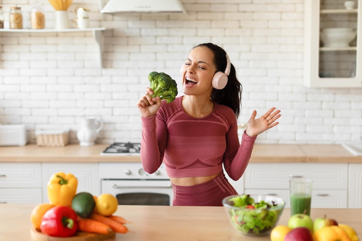 Zdjęcie szczęśliwa kobieta fitness w słuchawkach śpiewająca z brokułami w kuchni