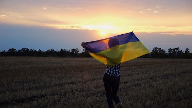 Zdjęcie szczęśliwa kobieta idzie po łące jęczmienia i odwraca się z podniesioną nad głową flagą ukrainy