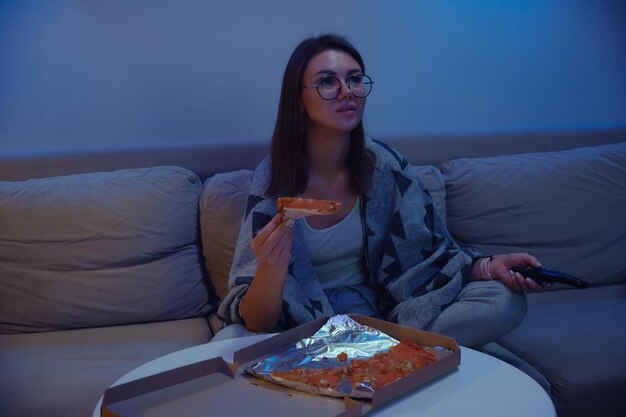 Szczęśliwa kobieta je pizzę w nocy oglądając telewizję, siedząc na kanapie w domu