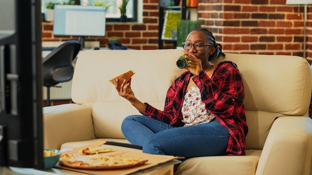 Szczęśliwa kobieta jedząca pizzę z dostawy do domu, oglądająca film w telewizji z butelkami piwa i fast foodami na wynos. Współczesna osoba dobrze się bawi, oglądając program w telewizji, na wynos.