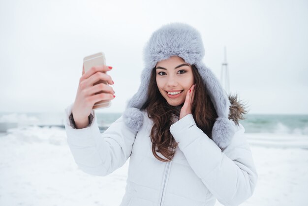 Szczęśliwa kobieta jest ubranym kapelusz robi selfie jej telefonem.