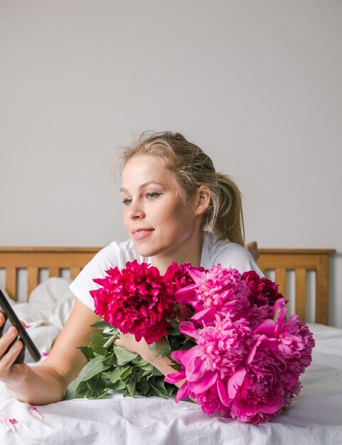 Szczęśliwa kobieta leżąca na łóżku w piżamie, ciesząca się bukietem tulipanów z piwonii i korzystająca z telefonu