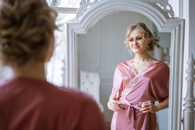 Szczęśliwa kobieta patrzy na swoje odbicie w lustrze w różowej jedwabnej szacie w pobliżu okna