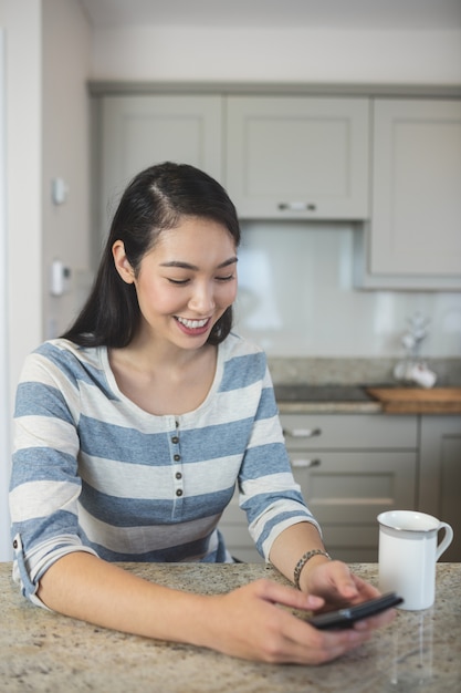 Szczęśliwa kobieta pisać na maszynie wiadomość tekstową na jej telefonie komórkowym w kuchni