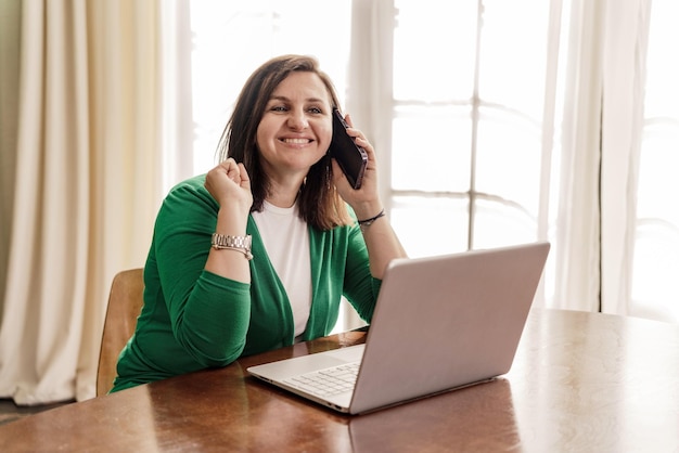 Szczęśliwa kobieta pracuje w domu online, odbiera telefon od klienta W pracy używa laptopa