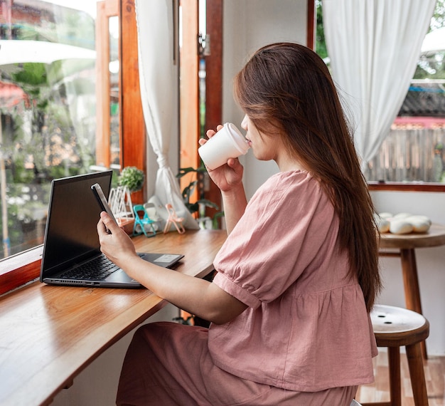 Szczęśliwa kobieta rasy mieszanej w kawiarni, korzystająca ze smartfona i laptopa, pijąca kawę w papierowym kubku