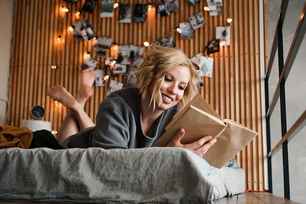 Szczęśliwa Kobieta Relaks I Czytanie Książki Na Wygodnym łóżku - Drewniana ściana I Zdjęcia Ze światłami - Niewyraźne Tło