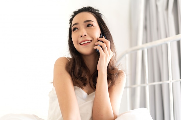 Szczęśliwa kobieta relaksuje używać i opowiada smartphone na łóżku w domu