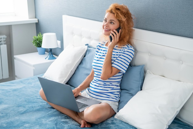 Szczęśliwa kobieta rozmawia przez telefon komórkowy w domu Dziewczyna podejmowania rozmowy telefonicznej gospodarstwa otwarty laptop na łóżku