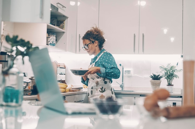 Szczęśliwa kobieta sama w domu gotuje lunch z uśmiechem Składniki i komputer online na pierwszym planie nieostre Koncepcja życia prawdziwej żony i samotnej kobiety Kobiety gotują w białej kuchni