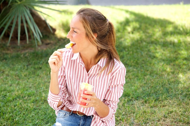 Szczęśliwa kobieta siedząca w parku i jedząca owoce.