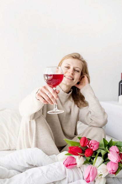 Szczęśliwa Kobieta Siedzi Na łóżku W Piżamie, Z Przyjemnością Delektując Się Kwiatami I Lampką Czerwonego Wina