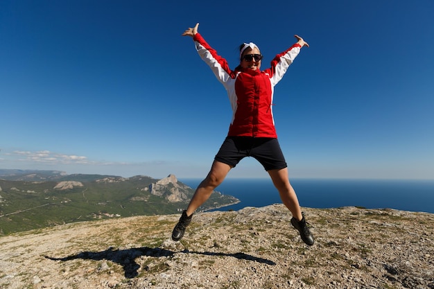 Szczęśliwa kobieta skacze na szczyt góry latem w górach, ciesząc się wspinaczką z pięknym skalistym i morskim tłem