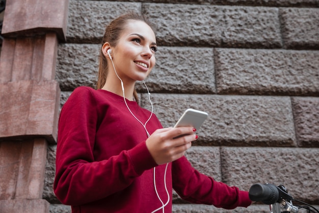 Szczęśliwa kobieta ubrana w sweter spacerująca z rowerem po mieście, stojąca przy ścianie i słuchająca muzyki przez telefon