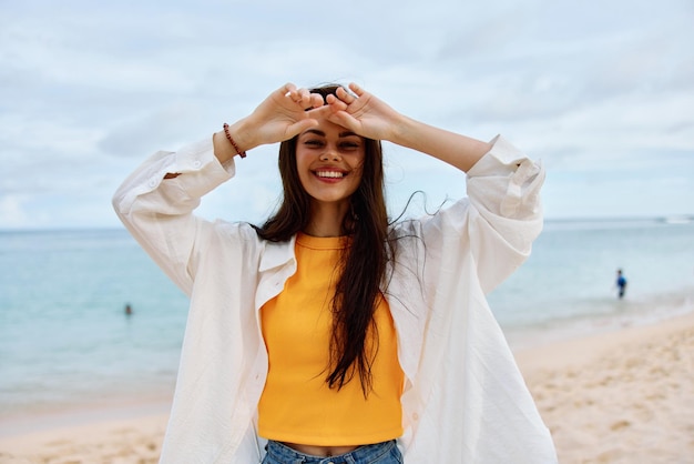 Szczęśliwa kobieta uśmiech z zębami z długimi włosami brunetka chodzi wzdłuż plaży w żółtej koszulce dżinsowych szortów i białej koszuli w pobliżu morza letnia podróż i poczucie równowagi wolności