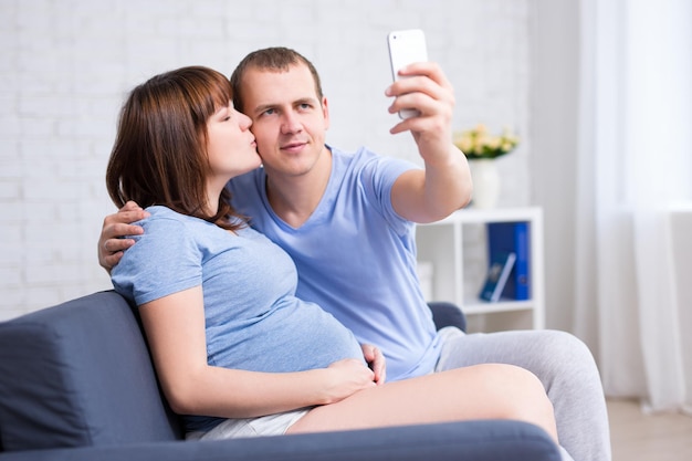 Szczęśliwa kobieta w ciąży całująca męża i robiąca zdjęcie selfie smartfonem w nowoczesnym salonie