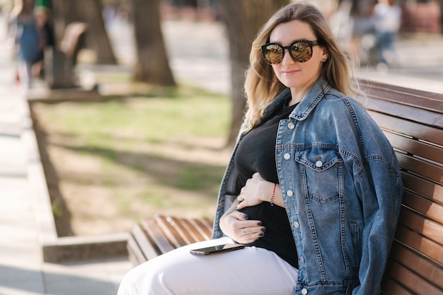 Szczęśliwa kobieta w ciąży kładzie ręce na brzuchu siedząc na ławce w parku