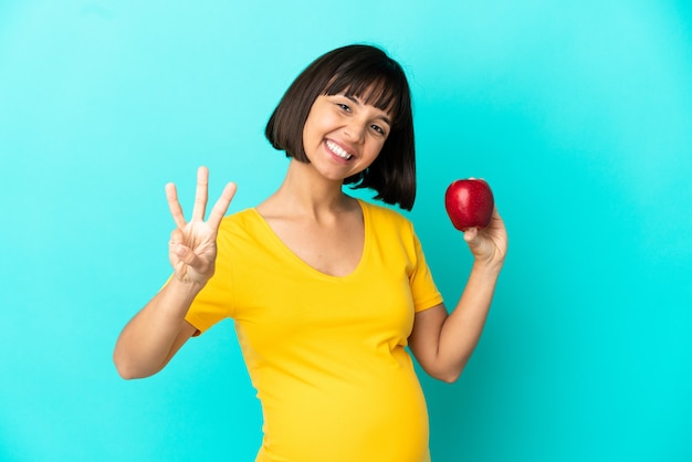 Szczęśliwa kobieta w ciąży trzymająca jabłko na białym tle na niebieskim tle i licząca trzy palcami