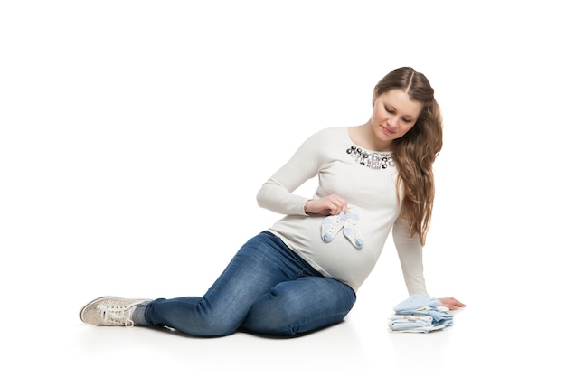 Szczęśliwa kobieta w ciąży trzymająca różne ubrania dla dzieci na białym tle