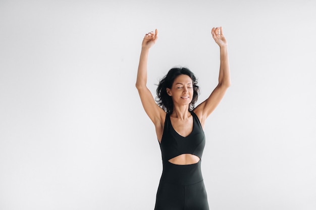 Szczęśliwa kobieta w czarnej odzieży sportowej stoi z rękami uniesionymi na jasnym tle Dziewczyna skacząca podniosła ręce do góry na siłowni