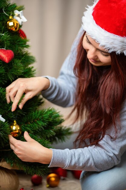 Zdjęcie szczęśliwa kobieta w kapeluszu św. mikołaja ozdabia choinkę noworoczną